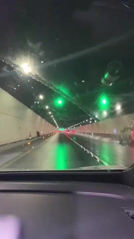 The video shows a view from inside a car driving through a tunnel. The tunnel has white walls with lights along the ceiling and sides. There are green lights visible further down the tunnel. The road surface appears wet, reflecting the lights. Other vehicles are visible ahead. The car's windshield has water droplets on it, and the interior of the car is partially visible in the foreground. The video captures the perspective of a driver or passenger moving through the tunnel.