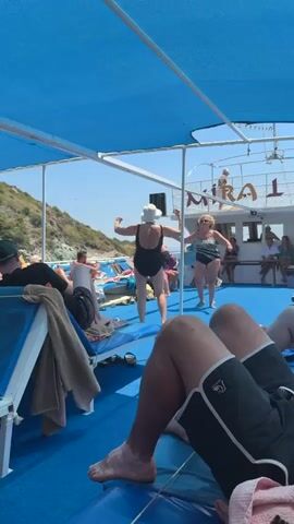 A video shows a group of people on a boat, most of them wearing swimsuits. The boat has blue seating and a blue canopy. Two women in swimsuits are dancing near the front of the boat. One woman is wearing a black swimsuit and a white hat. The other woman is wearing a black and white patterned swimsuit. There are other people sitting and standing around the boat. In the background, there is a body of water and a coastline with green hills. The sky is clear and blue. The word "MIRA" is visible on the side of the boat. The camera angle is from a seated position, looking towards the front of the boat.