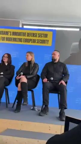 The video shows three people seated on chairs for a panel discussion. A woman in a black jacket and boots sits on the left, a woman in a black dress and jacket in the middle, and a man in all black on the right. The man in black is holding a microphone. A large screen behind them displays a woman on video call. A banner on the screen reads "UKRAINE'S INNOVATIVE DEFENSE SECTOR FOR MODERNIZING EUROPEAN SECURITY" and "UKRAINE HOUSE DAVOS" is repeated along the bottom. A laptop is partially visible in the bottom right corner.