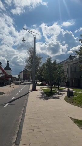 The video shows a street scene on a sunny day. The camera is looking down a paved road with a bike lane. To the right of the road is a sidewalk made of light-colored paving stones. A tall, gray lamppost with a decorative top and a single light fixture stands on the sidewalk. Several trees with green leaves are planted in a grassy area next to the sidewalk, casting shadows on the ground. In the background, there are buildings and a church tower with dark spires. There is construction equipment visible, including a crane. The sky is blue with fluffy white clouds. The sun is shining brightly, creating lens flare in the upper right corner of the video. There are no people visible in the video.