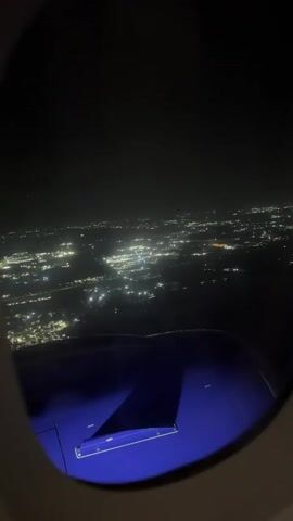 A view out of an airplane window at night. The lights of a city or town stretch across the horizon. The wing of the airplane is visible in the foreground, illuminated by blue light. The sky is dark.