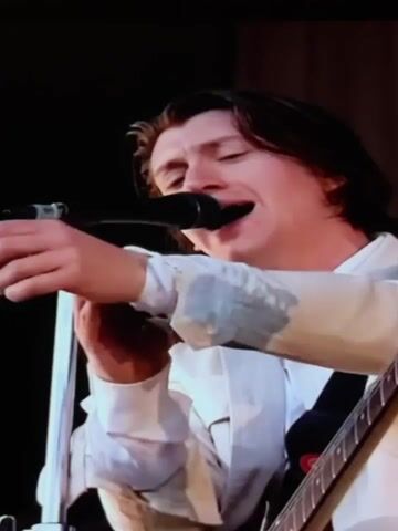This is a short video clip showing a musical performance on a stage. The primary subject is a male musician, likely a singer and guitarist, performing in front of an audience. He is wearing a white shirt and pants. He is seen singing into a microphone and playing a guitar. Another musician, possibly a drummer, is visible in the background. The stage has a dark backdrop and some lighting equipment. The overall mood appears energetic, typical of a live concert. There is no visible text in the video. Given the location context of Leninsk-Kuznetskiy, Russia, this could be a concert happening in that area. The time of day is not explicitly indicated, but the stage lighting suggests it could be evening or nighttime.