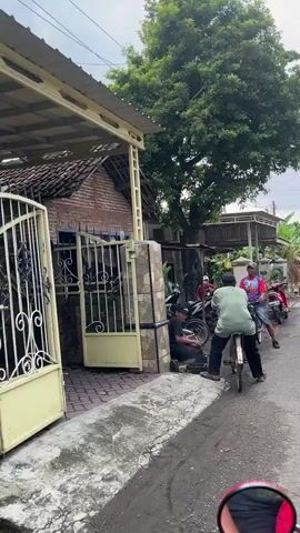 The video shows a street scene in front of a small building that appears to be a bicycle repair shop. Several people are present. In the foreground, a person is sitting on a bicycle facing away from the camera. To the left, a gate leads to a property with a brick house. Inside the open-fronted building, several bicycles and bicycle parts are visible, including wheels. One person is sitting inside the shop, and another is seen lying down on a red motorcycle parked outside. Further down the street, two more people are on bicycles, one of whom is riding towards the camera. The ground is wet, suggesting recent rain. The overall impression is of a busy, informal commercial area.