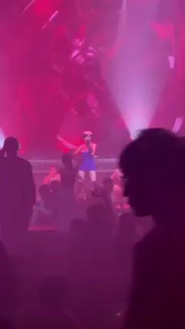 A person is recording a performance in front of a crowd. The stage is bathed in pink and white light. There is a backdrop that can be partially seen behind the performers.  Eight performers are on the stage, one in a blue dress and hat and the others in black outfits. They are dancing and the central performer seems to be singing. The crowd is silhouetted due to the stage lighting.