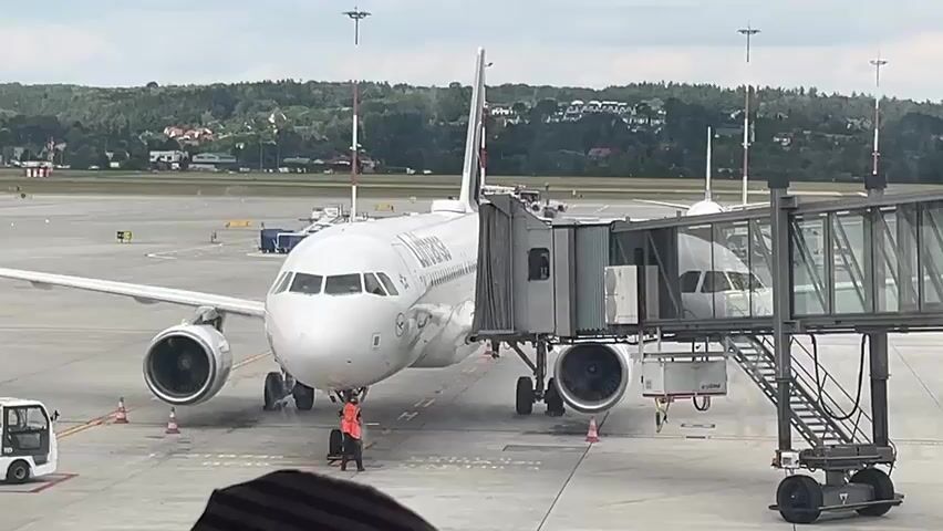 A Lufthansa airplane is parked at an airport gate, connected to a jet bridge. Two ground crew members are visible near the front landing gear of the aircraft. One crew member, wearing an orange vest, appears to be pushing a baggage cart. The scene is set on an airport tarmac under a cloudy sky, with trees and buildings visible in the background.