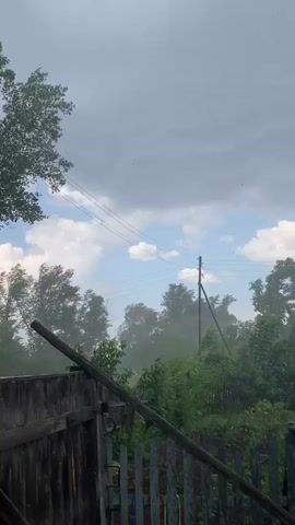 This is a video capturing a stormy scene in Rakity, Russia. The video begins by focusing on a wooden fence and gate, with lush green trees and foliage visible beyond. The sky is overcast with dark clouds and scattered patches of blue sky and white clouds, suggesting turbulent weather. A wooden utility pole with wires runs through the frame, adding to the rustic setting. The trees are swaying vigorously, indicating strong winds. The overall atmosphere is dramatic and suggests the onset of a storm or severe weather. There are no visible people or text within the video. The time of day appears to be daytime, given the brightness of the sky.