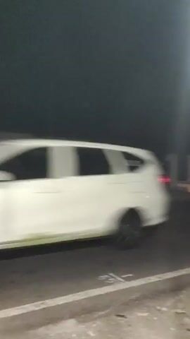 The video shows a dark exterior scene. A white fence is visible in the foreground, with a modern white house behind it. The house has several windows, some of which are illuminated. A bright street light is visible on the left side of the frame. The road in front of the fence is visible, with a yellow vehicle driving past. The vehicle is a four-door SUV with a spare tire mounted on the back. The video is slightly blurry and appears to have been taken from a moving vehicle.