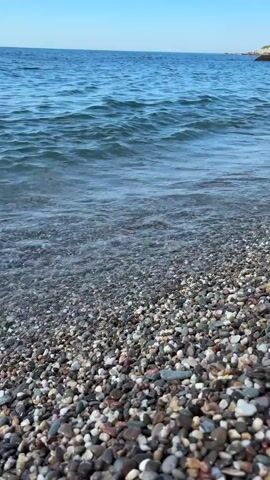 A close-up shot shows waves gently lapping against a pebble beach. The water is clear and blue, with small whitecaps. The pebbles are a variety of colors and sizes, from small, smooth stones to larger, rougher rocks. The background is a hazy blue sky. The sun is shining, and the scene is peaceful and serene.