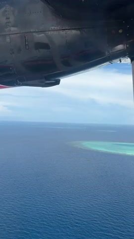 The video captures a view from an airplane flying over a vast expanse of deep blue ocean. The airplane's black and red fuselage is visible in the upper portion of the frame, along with a spinning propeller. Below the aircraft, the ocean stretches out to the horizon, where a cluster of small, turquoise islands or coral reefs can be seen. The sky is a clear blue with scattered white clouds. The overall impression is one of aerial travel over a tropical marine environment.
