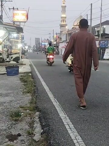 A person wearing a brown robe walks on the right side of the road, facing away from the camera. They are walking in the direction of traffic. A white car with its brake lights illuminated is in front of them, and a motorcycle with two people on it is to their left. In the background, there are buildings, a mosque with a tall minaret, and other vehicles on the road. The sky is overcast, and the overall lighting suggests it is either early morning or late afternoon. The road has a white line marking the edge.