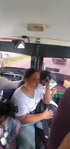 The video shows the interior of a bus. In the foreground, a woman with dark hair pulled back is seated, holding a smartphone to her ear and speaking. She is wearing a white t-shirt with blue stripes on the collar and a number "38" on the chest. To her left, a man in a white long-sleeved shirt is seen from the back, seated in the driver's seat and appearing to be operating the steering wheel. Further to the left and slightly behind him, another person is visible. In the lower left corner, a hand is holding up a smartphone, displaying a webpage. A child with dark curly hair, wearing a denim jacket over a pink garment, is seated to the left of the woman. Another person, mostly obscured by the foreground, is wearing a purple shirt and has long dark hair with a light purple accessory. The background shows a window with a view of a road and other vehicles, indicating the bus is in motion. The interior of the bus appears utilitarian, with visible lights and mirrors.