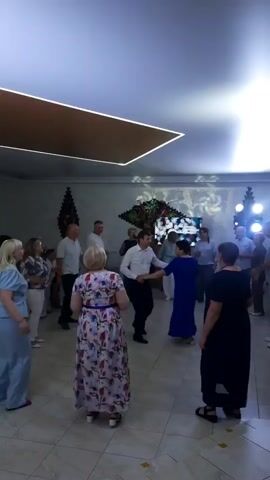 A group of people are dancing and socializing in a brightly lit room with a tiled floor. In the center, a couple is dancing, while others stand around watching. Some individuals are holding drinks, and the atmosphere appears festive. The room is decorated with floral arrangements and modern lighting fixtures.