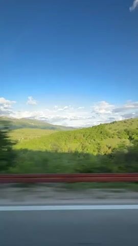 The video shows a view from a moving vehicle on a highway. The road is asphalt with white lines marking the lanes and a red guardrail separates the road from the adjacent landscape. The landscape consists of green hills, trees, and distant mountains, with a blue sky dotted with white clouds overhead. The camera is positioned inside the vehicle, looking out the side window. The video's main focus is the passing scenery.