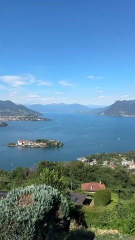 The video displays a scenic view of a large, calm body of water, likely a lake, under a clear blue sky with scattered white clouds. In the foreground, lush green trees and foliage are visible, along with a hint of a tiled roof and some shrubbery, suggesting a vantage point from a hillside or elevated area. Dominating the middle ground is a small island dotted with buildings, appearing to be a village or resort, surrounded by the water. Further in the distance, the expansive water stretches towards a range of majestic mountains that frame the horizon. The mountains exhibit varying shades of blue and gray, indicating depth and atmospheric perspective, with some settlements visible on their lower slopes. Small boats, including what appears to be a ferry or larger vessel, are scattered across the water, and some distant shorelines with buildings can be observed. The overall impression is one of tranquility and natural beauty, with a mix of human habitation and natural landscapes.