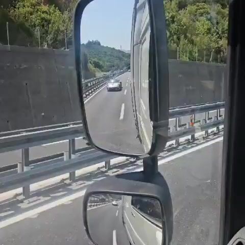 A silver Porsche is seen driving on a highway. The camera appears to be inside a truck, as evidenced by the presence of a truck's side mirror in the foreground of the first frame. The Porsche is in the right lane, and other vehicles are visible in the distance. The highway is bordered by a concrete barrier and lush green hills. The sky is clear and blue. The video shows the Porsche from various angles as the truck follows it.