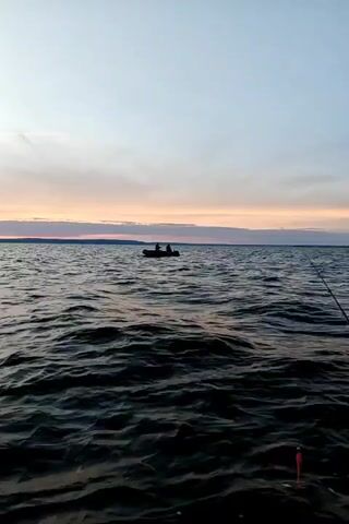 A static medium shot shows a small inflatable boat with two people fishing on a large body of water at dusk. The sky is a soft gradient of pale orange and pink, with wispy clouds. The water is dark and choppy, with visible waves. In the distance, a faint shoreline can be seen. A fishing rod is visible in the foreground on the right side of the frame. The two individuals in the boat are silhouetted against the sky. The boat appears to have a small motor attached to the back. The overall atmosphere is serene and tranquil.
