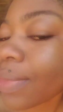 A woman with dark skin and dark hair braided on top, wearing a white shirt, is seen close up from the chest up, moving her face from side to side and smiling. Her left ear is visible in the frame at the beginning, and she has a mole above her upper lip.