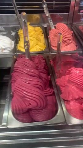 The video shows a close-up panning shot of a gelato display case filled with various flavors of gelato. The flavors visible include white, yellow, pink, dark red, light brown with cookies, yellow with caramel swirls, green with chocolate swirls, and dark brown. Each tub has a metal scoop inserted. The display case is made of stainless steel and appears to be in a commercial setting, likely an ice cream shop. The lighting is bright, illuminating the colorful gelatos.