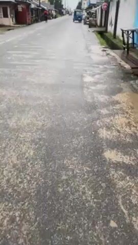 The video depicts a street after a rain. The road is paved with asphalt, which has a mottled texture. In the distance, buildings and other structures line the street. A small vehicle, possibly an auto-rickshaw, can be seen driving on the road. There are also pedestrians. The lighting suggests an overcast day. The overall tone is quiet and ordinary.