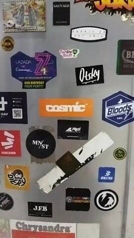 This is a close-up, vertical video of a collection of stickers on a metallic surface, possibly a refrigerator or locker. The stickers are varied in size, shape, and design, and appear to be from different brands and events. Some of the visible stickers include "Lazada," "7," "COSMIC," "MNST," "REI," "Bloods," "OTSKY," "JFB," and what appear to be brand logos and graphics. The metallic surface is somewhat reflective and shows some wear and tear, with a few smudges and scratches. There are no people or overt actions visible in the video; the focus is solely on the arrangement of the stickers.