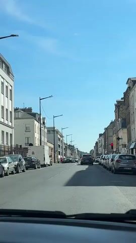 The video is shot from the perspective of a person inside a car, looking forward through the windshield. The car is driving down a street lined with buildings on both sides. The buildings appear to be residential or commercial, with multiple stories and windows. The street is paved, and there are parked cars along the right side of the road. The car is in motion, indicated by the slight blur of the passing scenery and the perspective of movement. The sky is blue with a few scattered clouds. There are streetlights and traffic lights visible along the street. A few other cars are on the road, and one car in front of the camera is clearly visible. There are shadows on the road, suggesting it is daytime. A person on a bicycle is visible on the left side of the road in the distance.