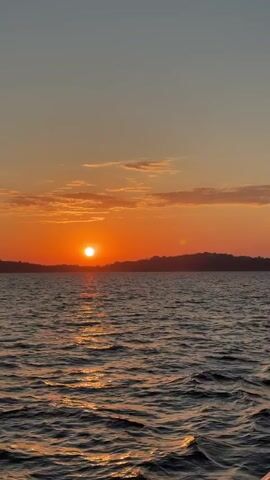The video shows a sunset over a body of water. The sun is a bright orange ball setting behind a dark treeline on the horizon. The sky above the sun is a gradient of orange and pink, with a few wispy clouds. The water is choppy and reflects the orange light of the setting sun. The camera appears to be on a moving boat. Someone speaks in the background.