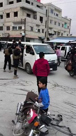 The video shows a street scene in what appears to be an urban area, possibly in the Middle East. There are several people present, including children and adults. Vehicles such as a white van, motorcycles, and a gold sedan are visible, navigating the cracked pavement. A group of individuals, possibly security forces, are gathered in the background, wearing dark clothing and carrying what appear to be firearms. People are walking around, and some are riding motorcycles. The overall atmosphere appears tense and busy.
