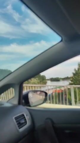 The video shows a view from inside a car traveling across a bridge over a river. The sky is partly cloudy and blue. The river is wide and appears calm. Trees line the riverbanks. In the distance, there are buildings and structures. A side mirror reflects the road ahead, showing other cars and a pedestrian walkway. The interior of the car is visible, including part of the dashboard and door panel. A road sign is partially visible on the left side of the screen. The camera is angled to capture the scenery outside the car and the reflection in the side mirror.