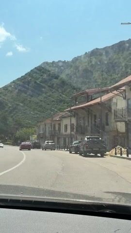 The video is shot from the perspective of someone inside a vehicle, looking forward through the windshield. The vehicle is driving on a paved road, with a white line marking the lane. On the right side of the road, there are several parked cars, including an SUV and a sedan. Beyond the parked cars, there are buildings with balconies and a mountain covered in vegetation rising in the background. The sky is blue with scattered white clouds. On the left side of the road, there are trees and more parked cars. The road curves to the left, with a red car in the distance ahead. The sun appears to be shining, casting some glare on the windshield.