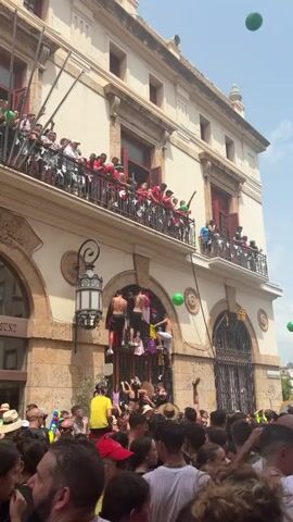 The video shows a crowd of people gathered in front of a building with a balcony and arched doorways. Several individuals are climbing on the building's facade, using hanging clothes and architectural features as support. Some people on the balcony are throwing green balloons down towards the crowd. The atmosphere appears to be festive, with many people in casual clothing and some wearing hats. The building has a light-colored stone facade and decorative lamps.