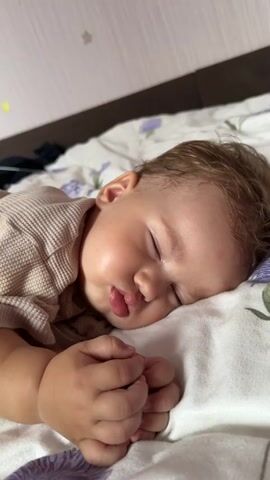 A baby is sleeping on its side. The baby is wearing a tan shirt with a waffle knit texture. The baby's head is resting on a white pillow with a floral pattern. The baby's hands are clasped together in front of its face. The background is a light pink wall.