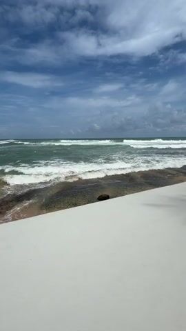 The video shows a beach scene with waves crashing on the shore. A white walkway runs along the edge of the beach, with palm trees providing shade. There are tables and chairs set up under umbrellas, suggesting a restaurant or bar area. The sky is blue with scattered clouds. There are no visible people or animals in the foreground of the video.