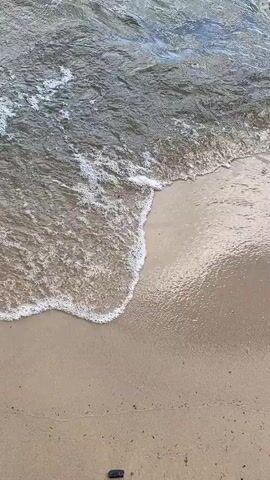 The video shows a close-up of gentle waves washing over a sandy beach. The water is clear and reflects the light, creating ripples and small bubbles as it recedes. The sand is light brown and appears wet near the waterline. There is a dark object, possibly a piece of debris or a shell, visible on the sand in the lower part of the frame. The waves are small and break softly, creating a calm and peaceful atmosphere.