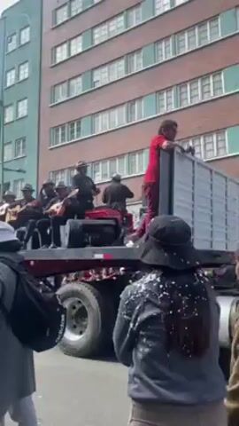 The video depicts a band performing atop the flatbed of a large truck moving along a city street. The band, consisting of several men, are seated and playing guitars, while one man sings into a microphone. They are all dressed in black, and most wear hats. The truck is moving at a slow pace, and a crowd of people surrounds it, some watching the performance and others walking alongside. Many people are wearing hats or have confetti on their clothing. A tall building with numerous windows stands in the background, the video is shaky, indicating that it was taken by someone moving with the crowd.