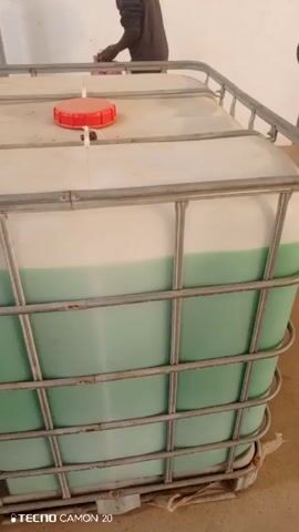 The video shows several IBC totes sitting on a light brown floor. Each tote is a square plastic container with a metal cage around it. The totes are filled with a light green liquid, leaving the top portion of the container empty. One tote has a red cap on top. The video pans from one tote to the others in a row. A person in dark clothing is briefly seen standing behind one of the totes.  The environment is a room with white or light-colored walls.  The video ends with the camera focused on a single tote.
