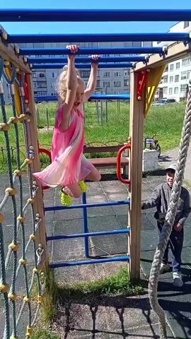 A young girl with blonde hair and a rainbow-colored dress hangs from monkey bars on a playground. She is wearing bright yellow Crocs. She is looking at the camera and smiling. A boy in a gray sweatshirt and blue pants stands to the right of the playground equipment, looking at the girl. The playground is made of metal and wood, with a rope net on the left side. The ground is covered in dark mulch. In the background, there is a grassy area, a building, and a blue sky. The sun is shining.