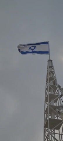 A low-angle shot captures the flag of Israel waving atop a white steel-framed tower against a mostly overcast sky. The flag, white with two horizontal blue stripes and a blue Star of David in the center, is positioned at the upper center of the frame. The tower, constructed of triangular support elements, rises from the bottom, slightly right of center. The sky varies in tone from gray to a lighter, almost white-tinged gray, suggesting the presence of sunlight behind the clouds. There is a barely audible ambient sound.
