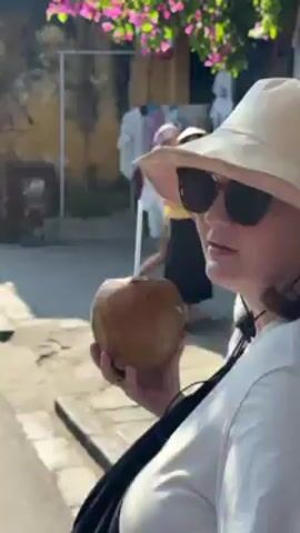 A woman wearing a white hat, white t-shirt, sunglasses, and a black bag across her body is holding out her hand as if to say “stop” to the camera. She then turns around and begins walking down a busy street lined with shops, holding a coconut with a straw. The street is decorated with Vietnamese flags. A man in a blue t-shirt and black shorts walks ahead of her. Other people are visible walking along the street. The video appears to be taken from a “follow me” perspective. The woman says "Follow me."