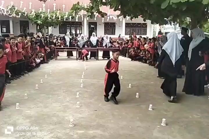 A group of children, dressed in red shirts and black pants, are participating in a game on a dirt ground in front of a building. The children are spaced out with small white cups placed on the ground in front of them. Two children are actively engaged in the game, running and interacting with the cups. Spectators, also in red shirts and black pants, are lined up on either side of the playing area, observing the game. Several adults, some wearing hijabs, are also present, watching the children. The environment appears to be outdoors, with trees and a building in the background. The scene is captured with an OPPO A96 phone, timestamped August 14, 2026, at 09:26.