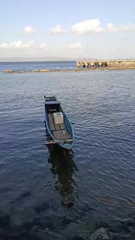 A blue wooden boat is anchored in calm water. The boat contains several white boxes and wooden planks. In the background, a pier extends into the water, and there is a rocky shore in the foreground. The sky is blue with scattered clouds.