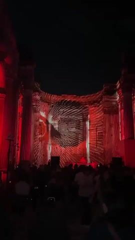 A crowd of people are gathered outdoors at night, watching a large projection on a building facade. The projection displays abstract, red-toned visuals, including an eye, geometric patterns, and what appears to be a play button. The building has large columns and architectural details illuminated by red lights. A DJ or performer is visible on a raised platform in front of the projection. The atmosphere seems to be that of an outdoor event or concert.