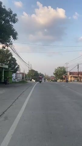 This is a vertical video shot from the perspective of someone on a moving motorcycle on a paved road. The road has a white dividing line on the left side. The sky is blue with scattered white clouds. There are utility poles and wires running parallel to the road on both sides. Buildings and trees line the sides of the road. Several other motorcycles are visible on the road, some ahead and some passing by. The rider in the foreground wears a blue shirt and a helmet. The overall atmosphere suggests a daytime scene on a street in a populated area.