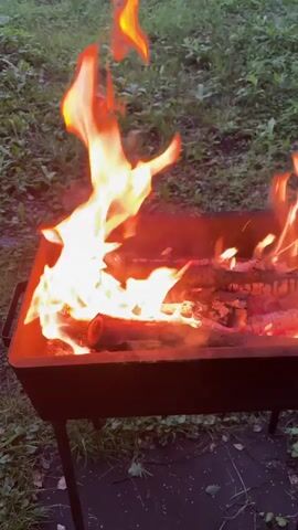 A video shows a fire burning in a dark metal grill outdoors. The grill has four thin black legs and is positioned on a surface covered with black fabric, surrounded by green grass and fallen leaves. The fire is vigorous, with bright orange and yellow flames engulfing logs of wood. Some smoke is visible rising from the fire. The video is shot from a slightly elevated perspective, looking down at the grill. The lighting suggests it might be dusk or an overcast day, as the flames are the dominant light source.