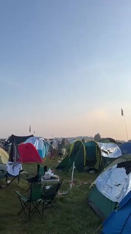 The video pans across a large campsite filled with numerous tents of various colors and sizes. The sky is clear with a few wispy clouds, and the sun is rising, casting a soft, warm light. Several folding chairs are set up near some of the tents, and a small table with some items on it is visible in the foreground. Laundry is hung to dry on lines between some of the tents. The overall impression is that of a campground at dawn.