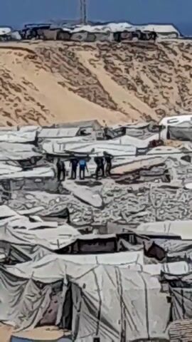 A wide shot shows a densely packed tent city in a desert environment. In the foreground, rows of white tents are visible. In the middle ground, a group of approximately five people are gathered amidst rubble, with several tents appearing damaged or collapsed. The people appear to be wearing dark clothing and one person is wearing a blue shirt. In the background, more tents are visible on a sandy hillside, with a tall structure, possibly a communication tower, rising behind them. The sky is clear and blue. The overall scene suggests a disaster or conflict zone where people are living in temporary shelters.