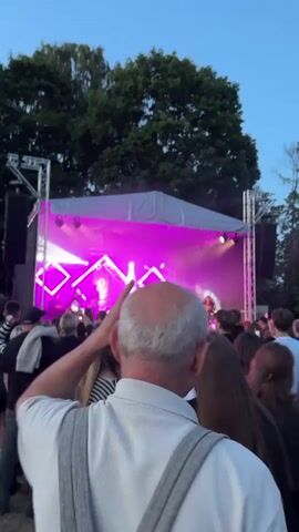 A concert is taking place outdoors at dusk. The stage is illuminated with pink and white lights, and a band is performing. A large crowd of people are watching the performance, and some are recording it with their phones. An elderly man in the foreground is facing away from the camera and appears to be enjoying the show. Trees and a clear sky are visible in the background.