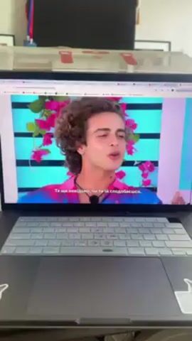 The video shows a laptop with a video playing on the screen. The video appears to be of a young man with curly hair speaking. Subtitles are visible in another language. The laptop is on a desk covered in snack food, including chips, a can of Corona Extra beer, and boxes of chocolates. A framed picture is in the background. The room appears to be indoors and lit by natural light. The laptop has a cat sticker on the bottom right corner. The camera pans across the table to show the variety of snacks.