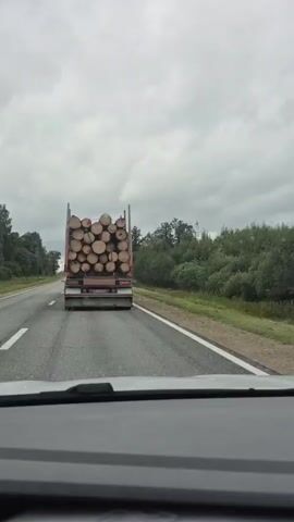 The video shows a truck loaded with logs driving on a two-lane road under a cloudy sky. The perspective is from inside a car following the truck. The truck is in the right lane. The road is surrounded by trees and vegetation.