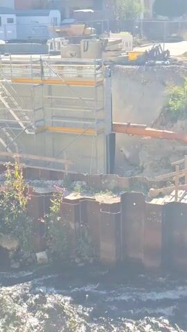 The video shows a construction site with a river in the foreground. The river has choppy water and some vegetation on its banks. The construction site has scaffolding, metal sheeting, and building materials. There are some buildings in the background. The video pans from right to left across the scene.