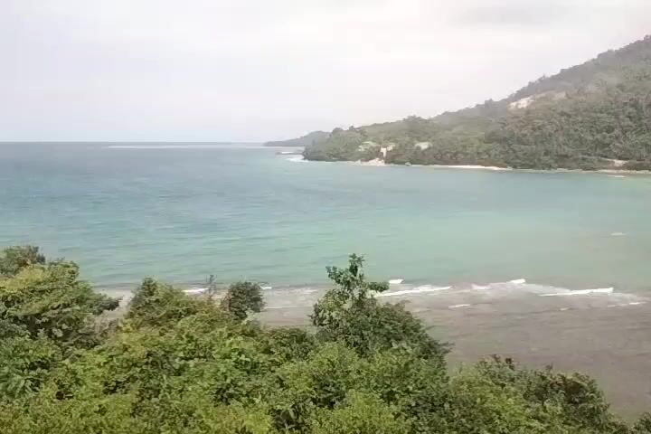 The video shows a scenic view of a bay with turquoise waters lapping at a sandy shore. Lush green trees and hills surround the bay, with a cloudy sky overhead. The camera angle is from a slightly elevated position, looking out over the water. There are no people or obvious man-made structures visible in the immediate foreground, though some structures can be seen nestled amongst the trees on the far side of the bay. The waves are gentle, creating a serene and tranquil atmosphere.